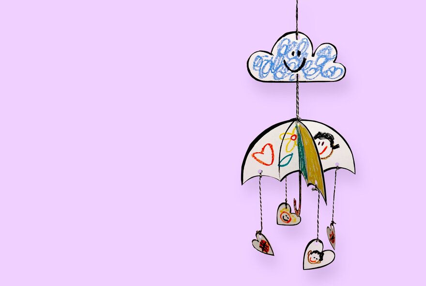 A hanging mobile of an umbrella with childish drawings on it and hanging from it in hearts, plus a blue swirly cloud above it; pink background.