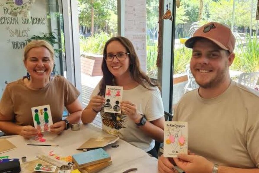Three smiling workshop participants seated at a table with craft materials, holding up jewellery they have made.