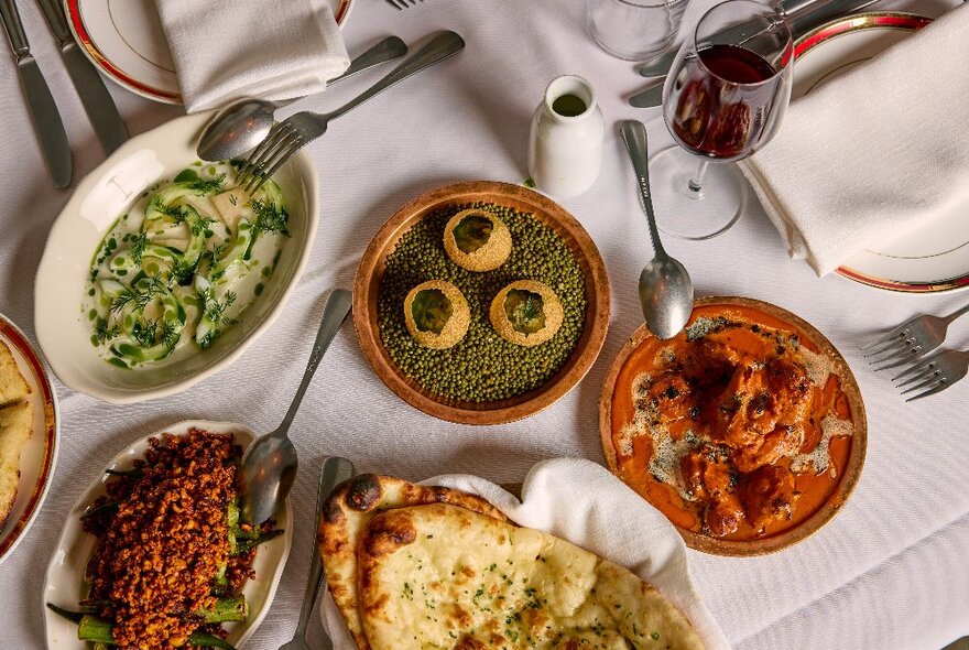 Looking down at a restaurant table at platters of Indian food, with white linen napkins folded atop of dinner plates.