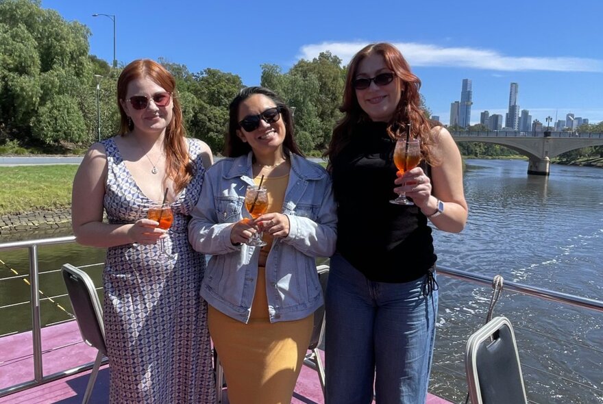 Three friends smiling and holding drinks aboard a brunch cruise boat with the Yarra River and Melbourne skyline in the background.