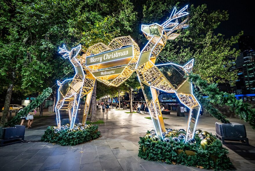 A glowing arch of two reindeer joined by a gold heart with 'Merry Christmas Melbourne' text, surrounded by green foliage.