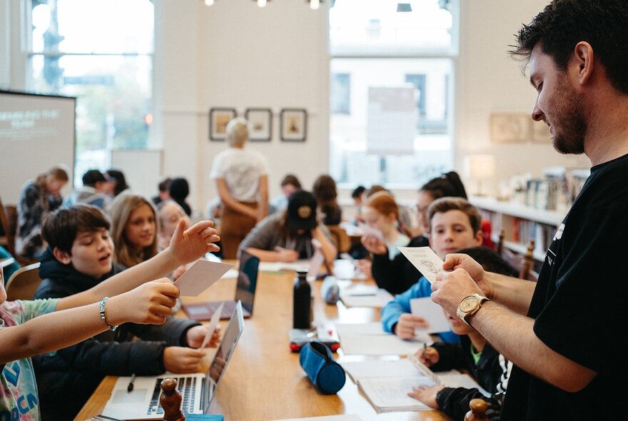 A teacher with paper in front of a table of children with hands raised.