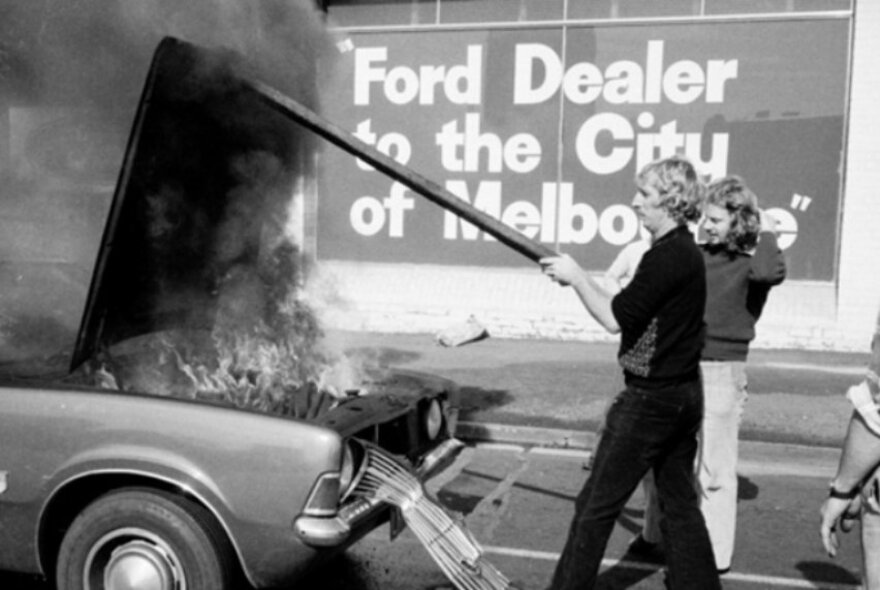 Black and white photo of people trying to extinguish a car's engine on fire, with Ford dealership sign in the background.