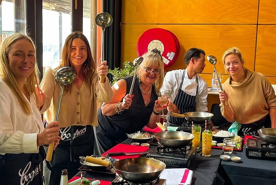 A group of people holding up ladles in front of small woks and cooking utensils, at a Thai cooking workshop that is taking place inside a restaurant .