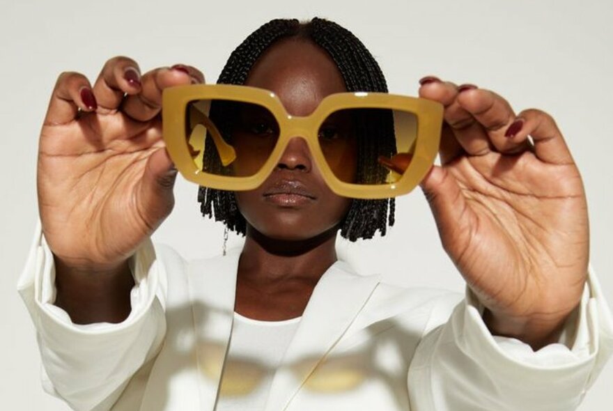 Model wearing a white suit holds up pair of sunglasses to camera