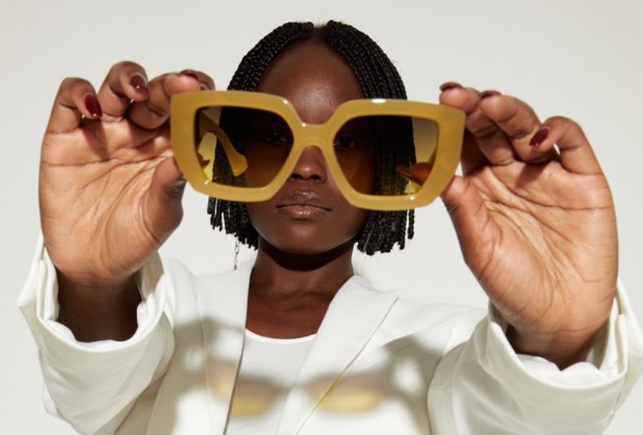 Model wearing a white suit holds up pair of sunglasses to camera