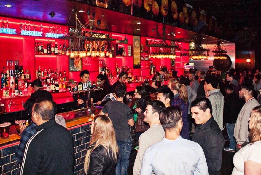 People milling around a bar that's illuminated with red lights. 