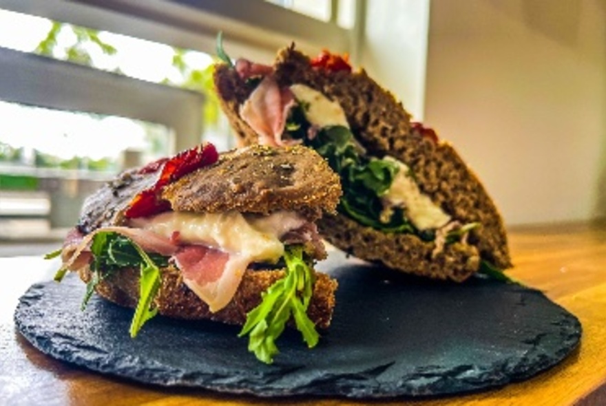 Plate on a cafe table displaying a soft bread roll cut in half and filled with lettuce, melted cheese and cured meat.