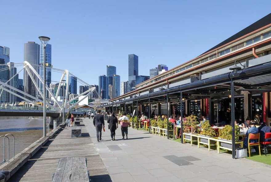 View of the exterior seating area of BangPop restaurant's outdoor terrace, with people walking along the wharf beside the river, and Melbourne's skyscrapers in the background.