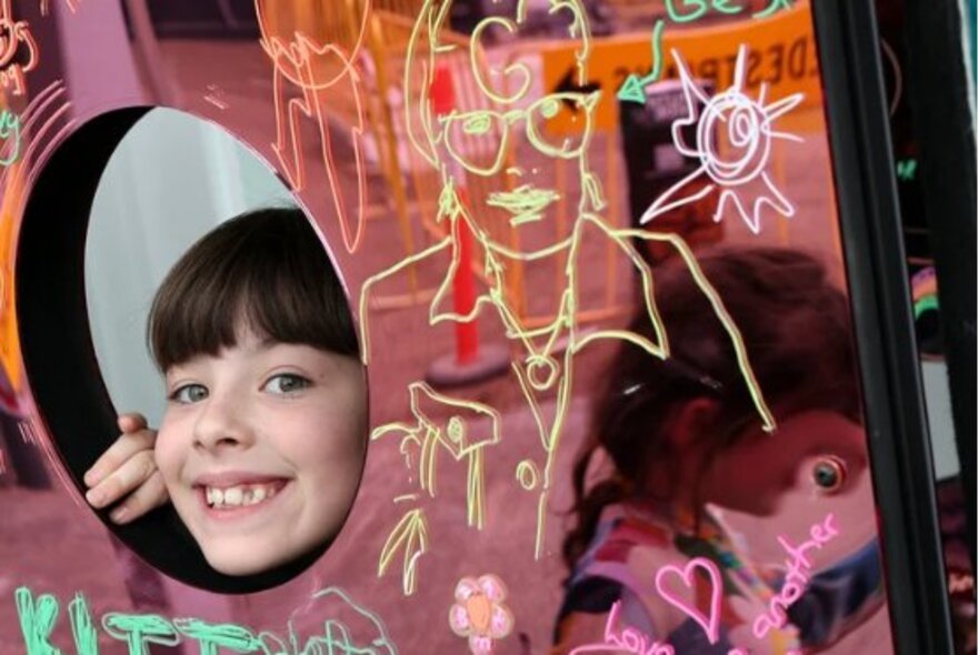 A smiling child poking their head through a round hole in a reflective scribble wall at the RESET Festival at Fed Square.