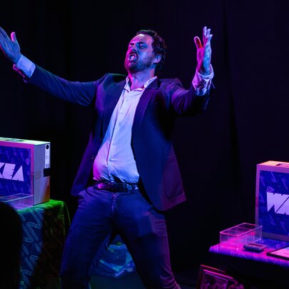 Performer on a darkened stage during a performance of the live comedy game show IKEA Wars, with two boxes on stage, one labelled IKEA and the other labelled WARS.