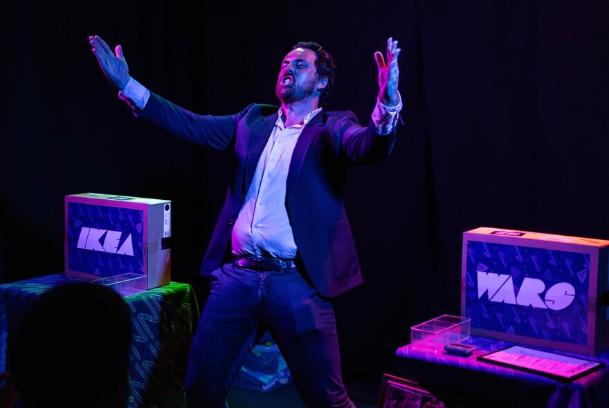Performer on a darkened stage during a performance of the live comedy game show IKEA Wars, with two boxes on stage, one labelled IKEA and the other labelled WARS.