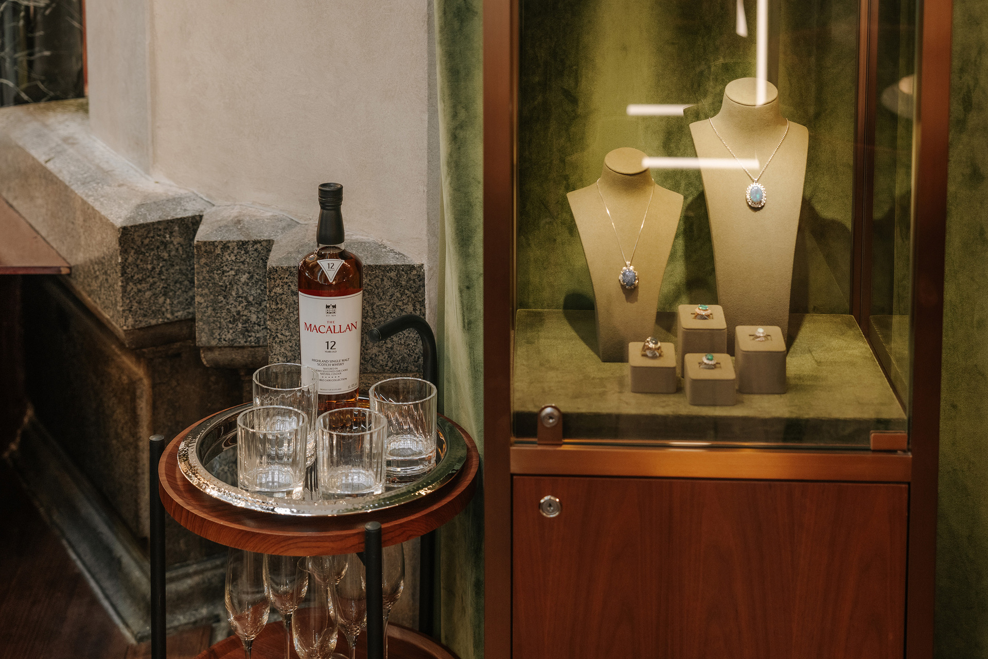 A display case at a jewellery store lined in green velvet there is a small table with a bottle of whisky an glasses next to it.