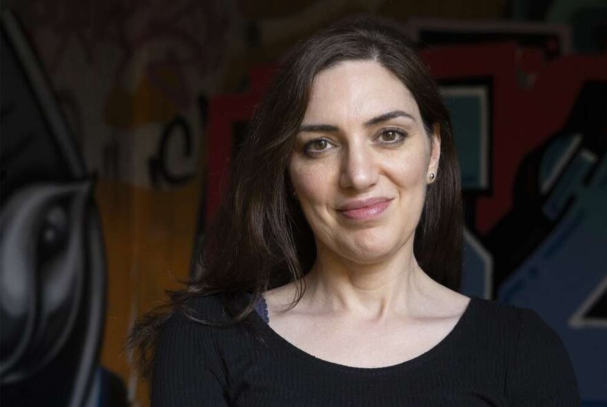 A woman with long dark hair smiling warmly against a dark, graffitied background. 