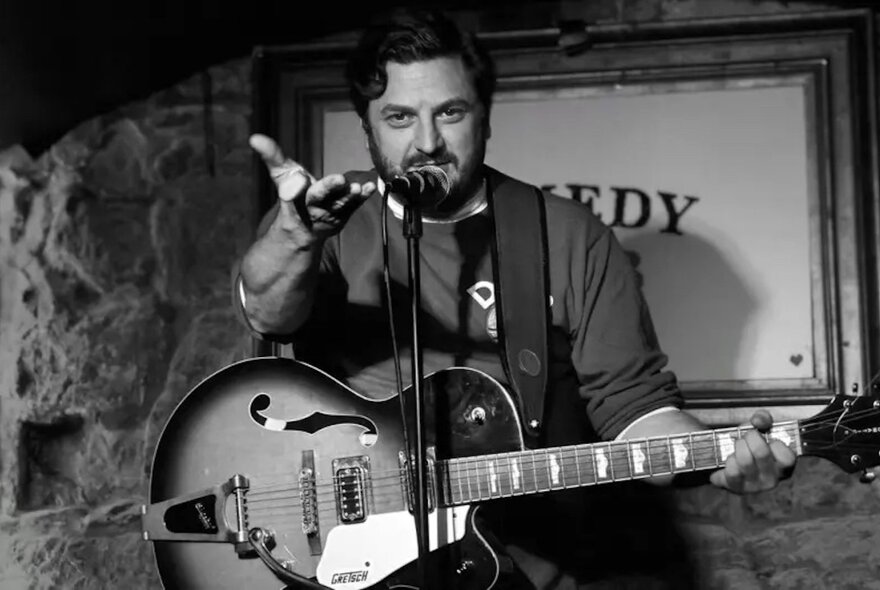 A black and white image of comedian George Rigden with a guitar talking into a microphone with a hand outstretched, palm upwards. 