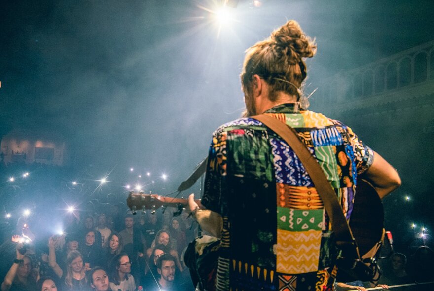 View of the back of singer Jeremy Loops, he wears a colorful patterned shirt and is playing an acoustic guitar in front of an audience in a medium sized venue. 