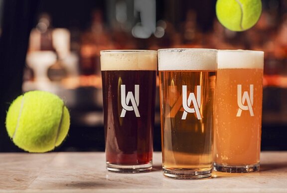 Three schooners of beer on a tabletop with two tennis balls mid-air alongside them.