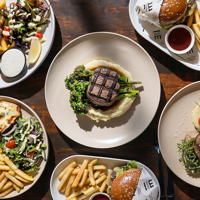 Looking down at plates of food that include steaks, burgers, salads and fries on a wooden restaurant dining table.