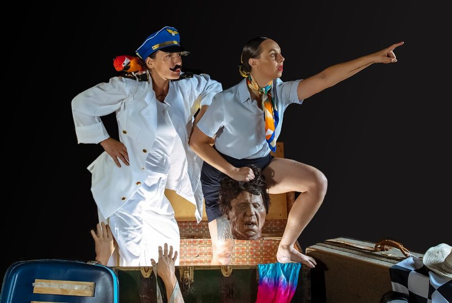 People dressed as cabin crew, one pointing into the sky while the other looks on with a head near their legs.