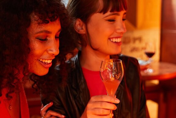 Two people are smiling and toasting with glasses of champagne at a dim, warm-lit bar.