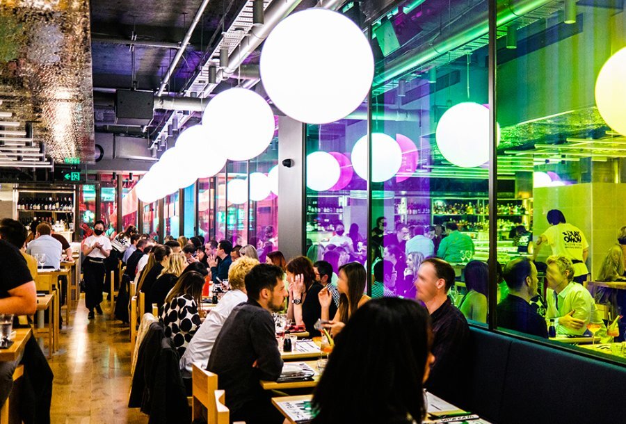 Diners in a restaurant with neon windows and large pendant lights.