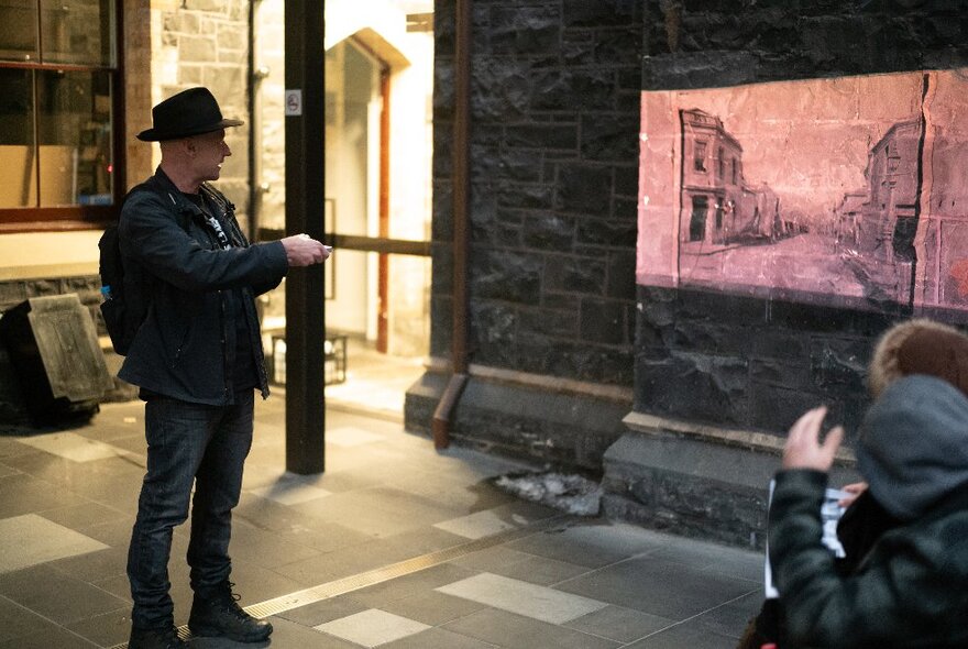 A man wearing a black hat and leading a walking tour, shining a projection from a hand held projector, onto a wall of a building in a city street.