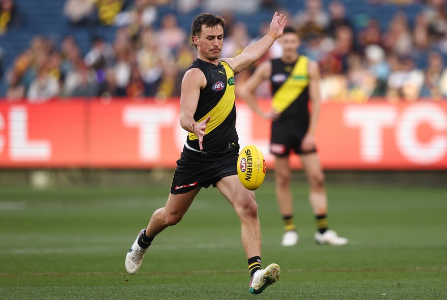 A Richmond AFL player running while about to kick a yellow football he has just dropped in front of his leg, with blurred fans in the stadium in the background.
