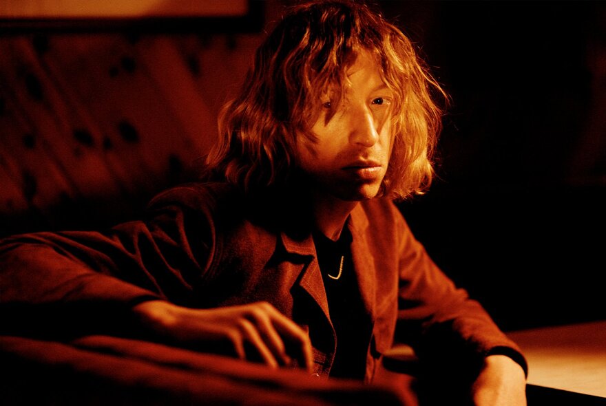 English electronic music producer Daniel Avery, with shaggy tousled hair half covering his eyes, looking at the camera, wearing a jacket and seated at a table.