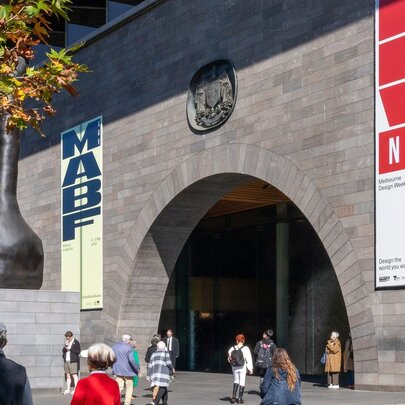 The grey concrete facade of the National Gallery of Victoria International building on St Kilda Road with banners advertising Melbourne Design Week and the Melbourne Art Book Fair, and people walking past.