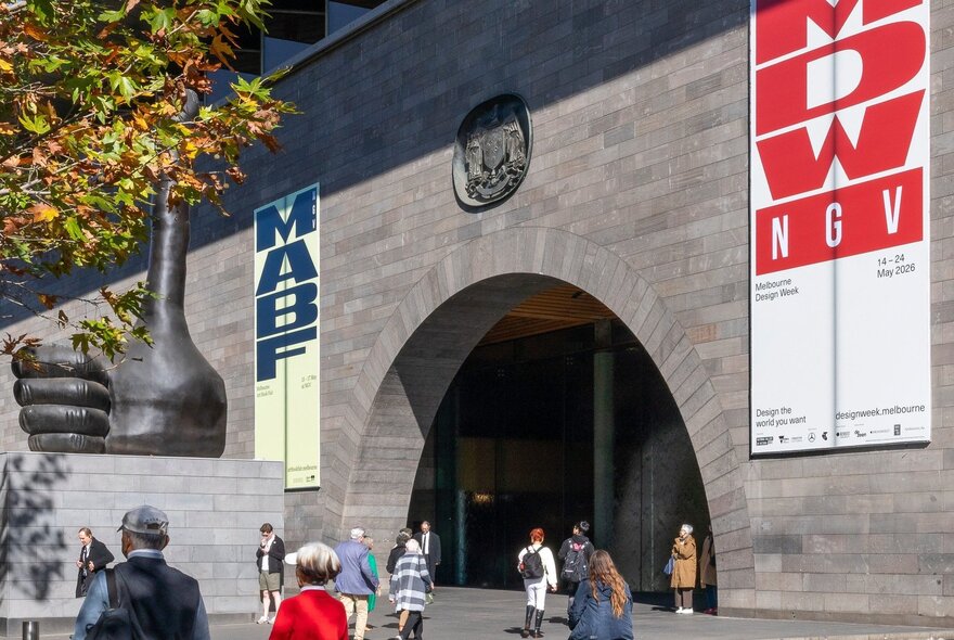 The grey concrete facade of the National Gallery of Victoria International building on St Kilda Road with banners advertising Melbourne Design Week and the Melbourne Art Book Fair, and people walking past.