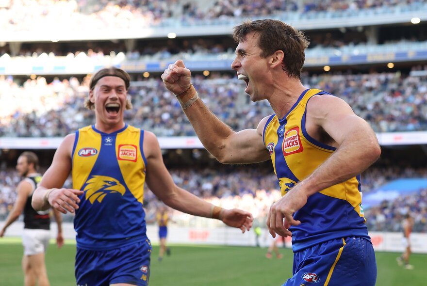 Two West Coast AFL players celebrating on the field,, with blurred fans in the stadium in the background.