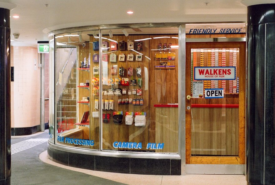 A curved glass display of a storefront in a basement arcade, with a light wooden door frame and a sign on the glass door reading 'WALKENS' in red capital letters.