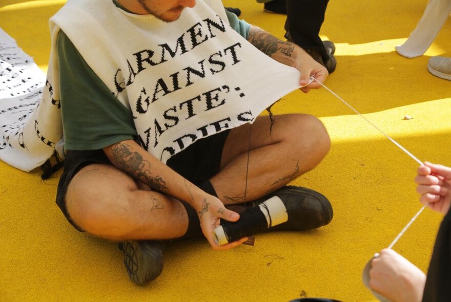 A man sitting cross-legged on a yellow floor while someone pulls a string from the bib he is wearing, unravelling the garment. 