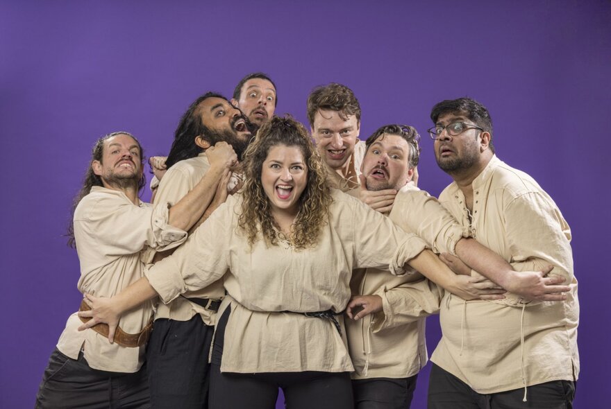 A comedy troupe all wearing beige tunics and black pants as six of them are held back by their only female member.