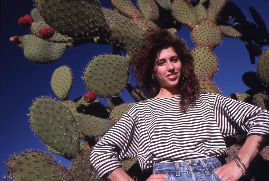 A photograph of a young woman in a black and white striped t-shirt, smiling down at the camera as she stands, hands on hips, in front of a large prickly pear cactus.
