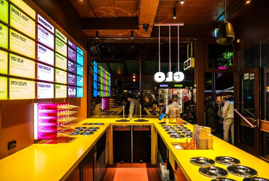 The interior of GLO Gelato with glowing menu board, a yellow counter top and people gathering outside. 