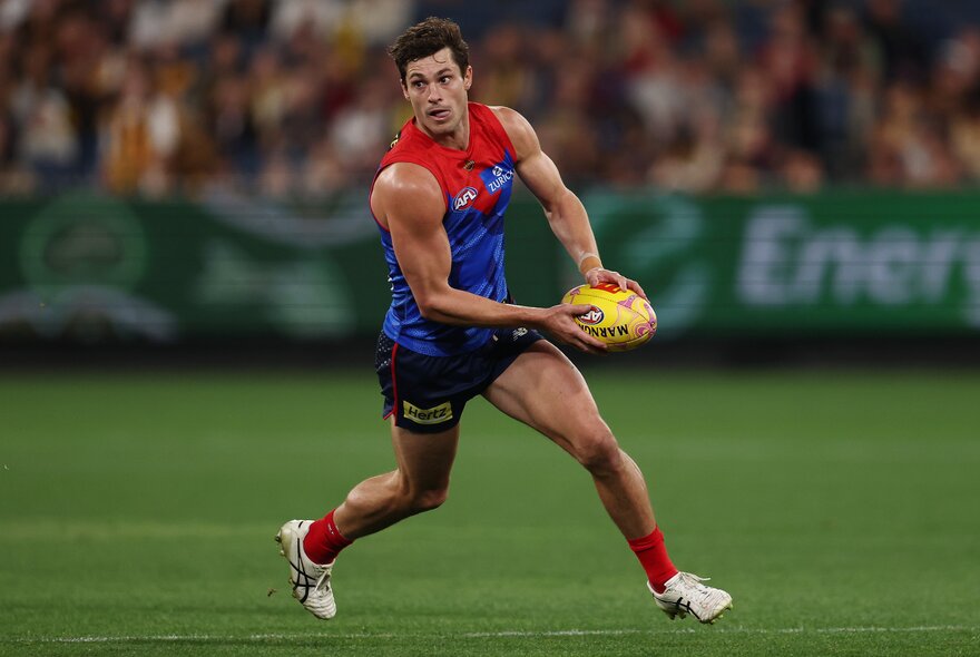 A Melbourne AFL player running while about to kick a yellow football he holds in his hands, with blurred fans in the stadium in the background.