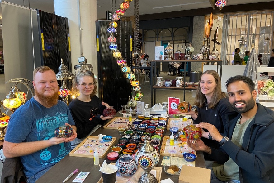 Four people seated at a workshop table in a shop setting, with rows of dishes and other craft tools.