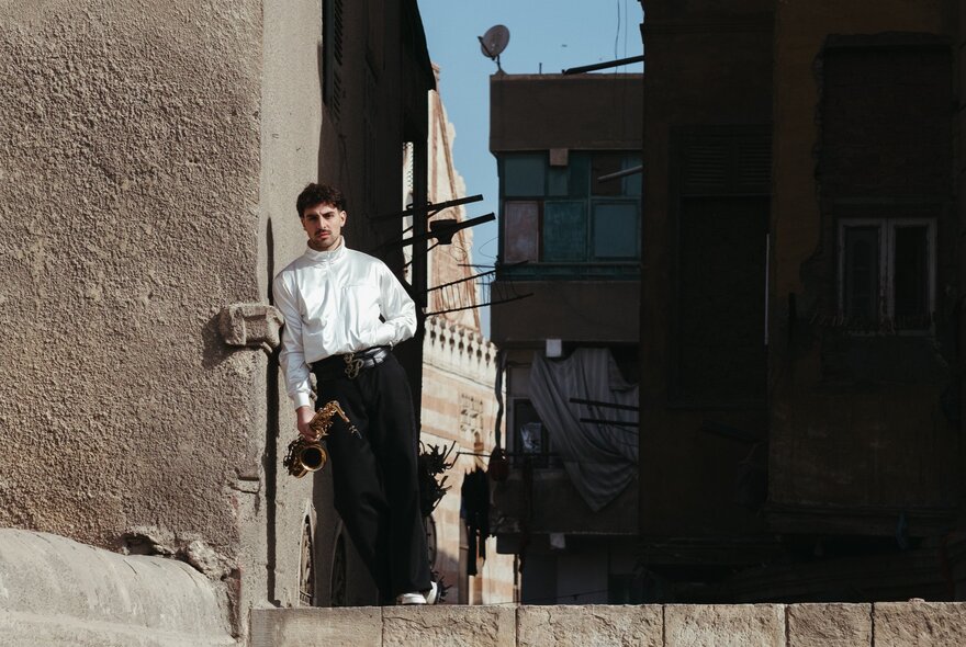 The musical artist Saint Levant, wearing dark trousers and a white shirt holding a saxophone by his side, leaning against a wall, with buildings in the shadows behind him.