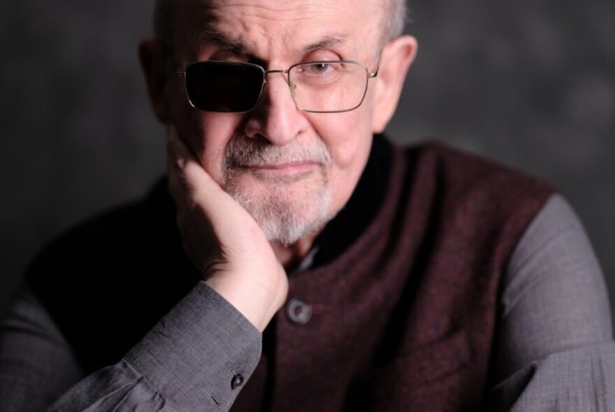 Portrait of Sir Salman Rushdie with his hand to his chin, wearing glasses, an aubergine vest and grey shirt.
