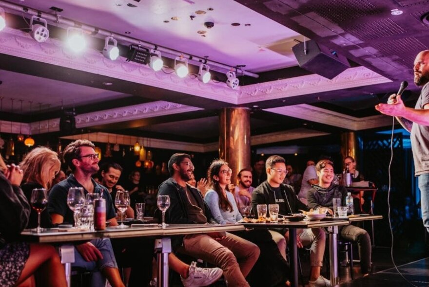 An audience seated in a comedy venue at tables with drinks in front of them, watching a stand-up comedian perform on stage.