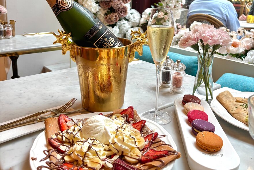 A brunch table setting with a plate of strawberry chocolate crepes with ice cream, a platter of  macarons, a flute of champagne, and a bottle of champagne resting in a gold ice bucket on a marble tabletop.