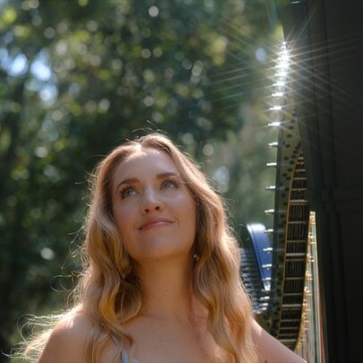 Harpist Emily Grainger outdoors in a garden with tall trees and sunlight hitting her harp. 