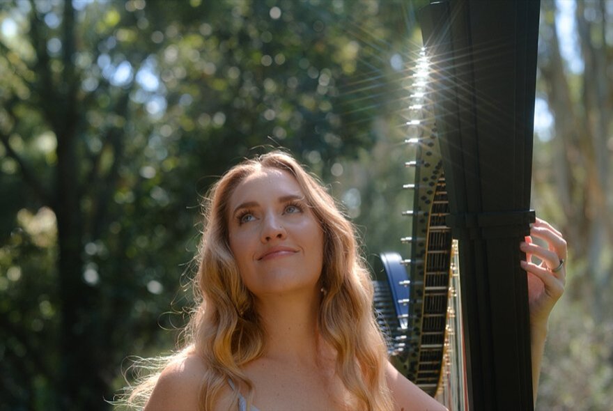 Harpist Emily Grainger outdoors in a garden with tall trees and sunlight hitting her harp. 