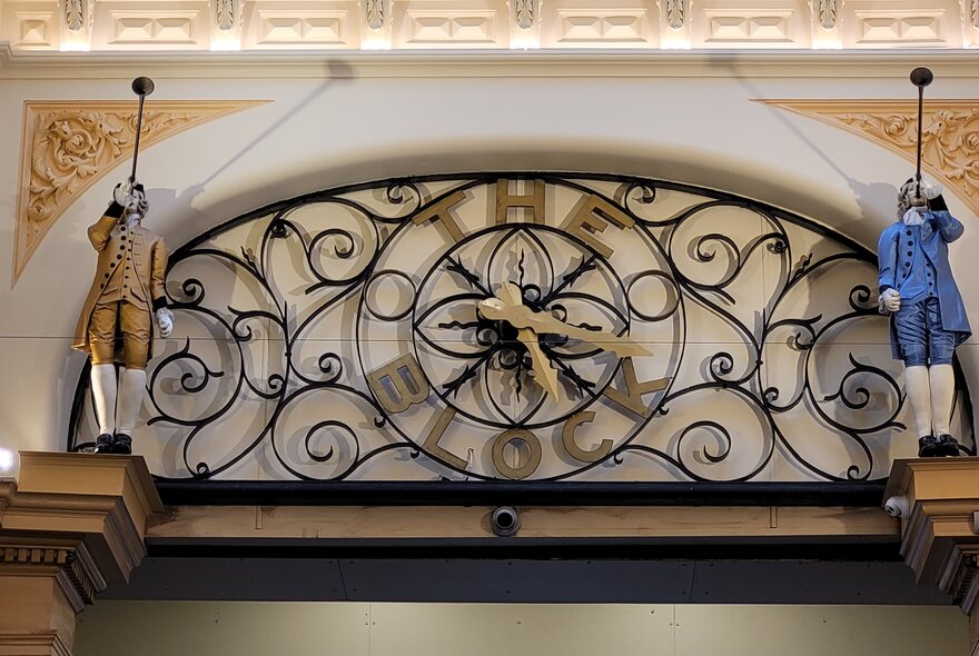 A wrought iron clock at the entrance to The Block arcade, with two figurines either side of it.