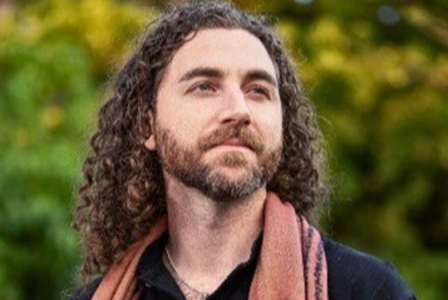 Man with his head on a slight angle, looking upwards, with curly hair and a beard, posed outdoors against greenery. 