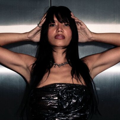 DJ and producer Zalina, her hands resting on either side of her head, wearing a strapless top, her long dark hair falling past her shoulders.
