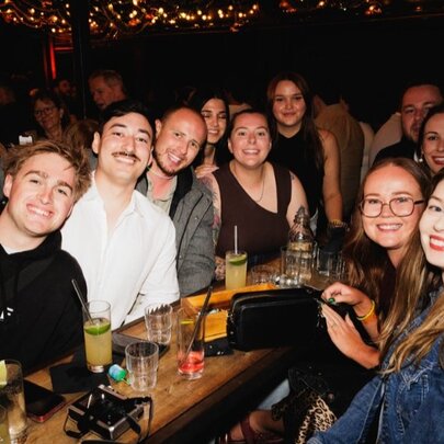 A large group of friends at a bar all around a single table with drinks and looking happy. 