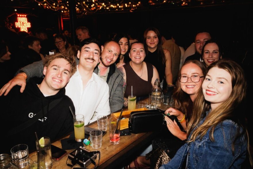 A large group of friends at a bar all around a single table with drinks and looking happy. 