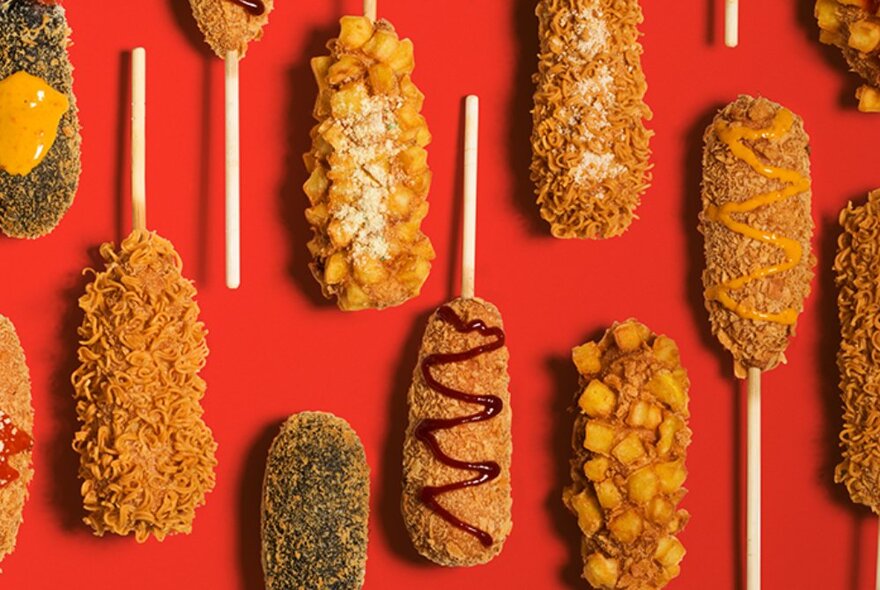 Crumbed and deep fried hot dogs on sticks, arranged on a red background, with different sauce toppings.